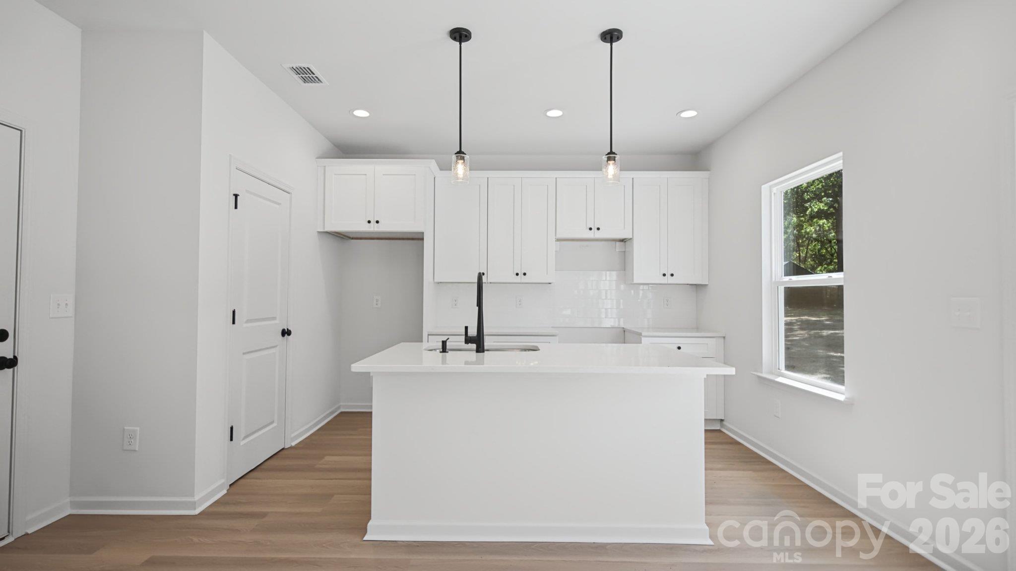 Bright white kitchen with a large island, black faucet, subway-tile backsplash, and light wood flooring.