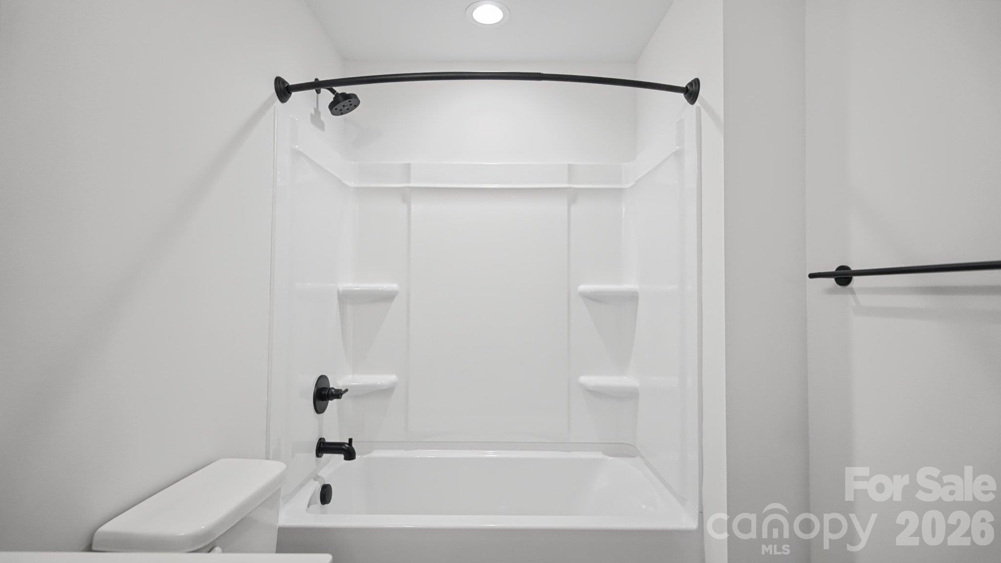 White tub and shower with molded shelves and matte black fixtures beside a partial view of the toilet.