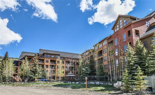 Multi-story condominium building with brick and timber finishes, framed by evergreen trees.