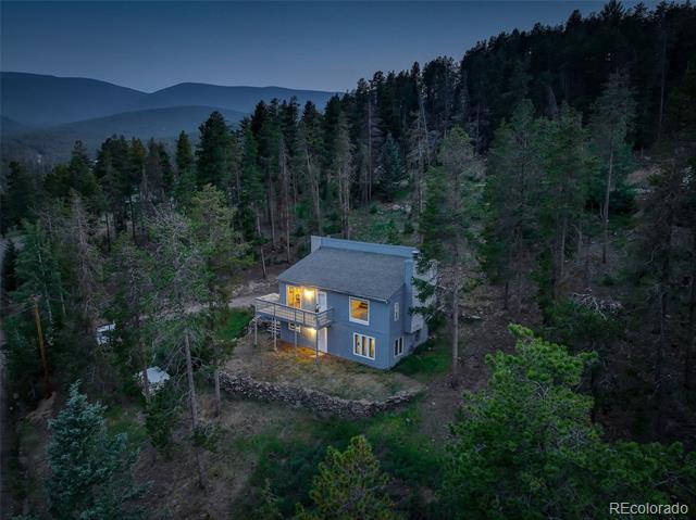 Aerial view of a blue-gray house with a raised deck among trees, mountain ridges beyond.