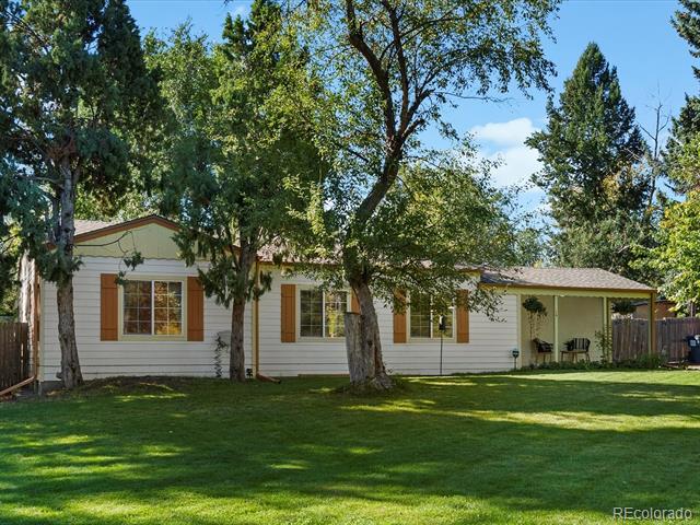 Single-story white home with brown shutters and a covered porch beneath mature trees.