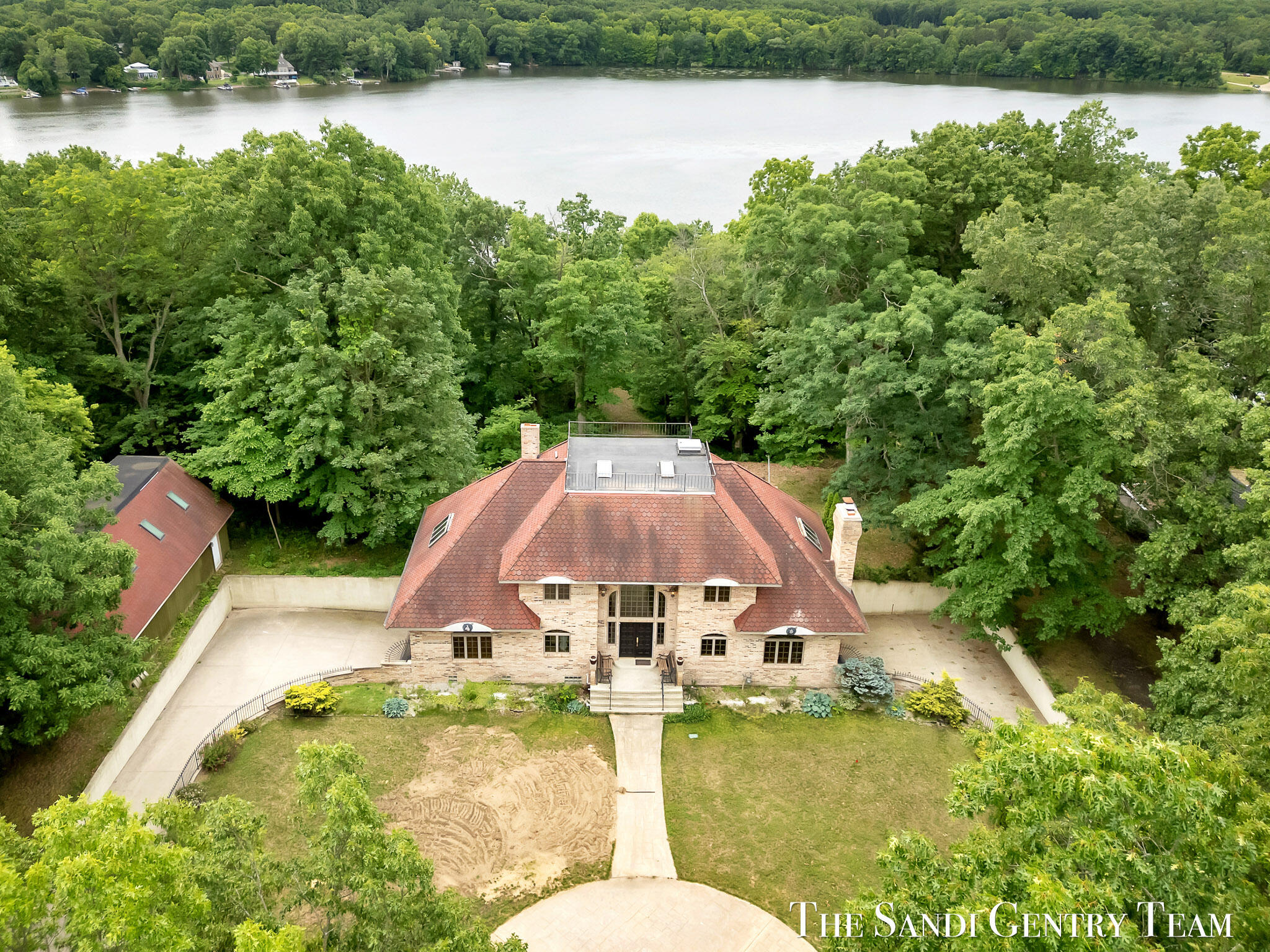 Aerial view of a stone house with a red tiled roof, surrounded by trees beside a lake.
