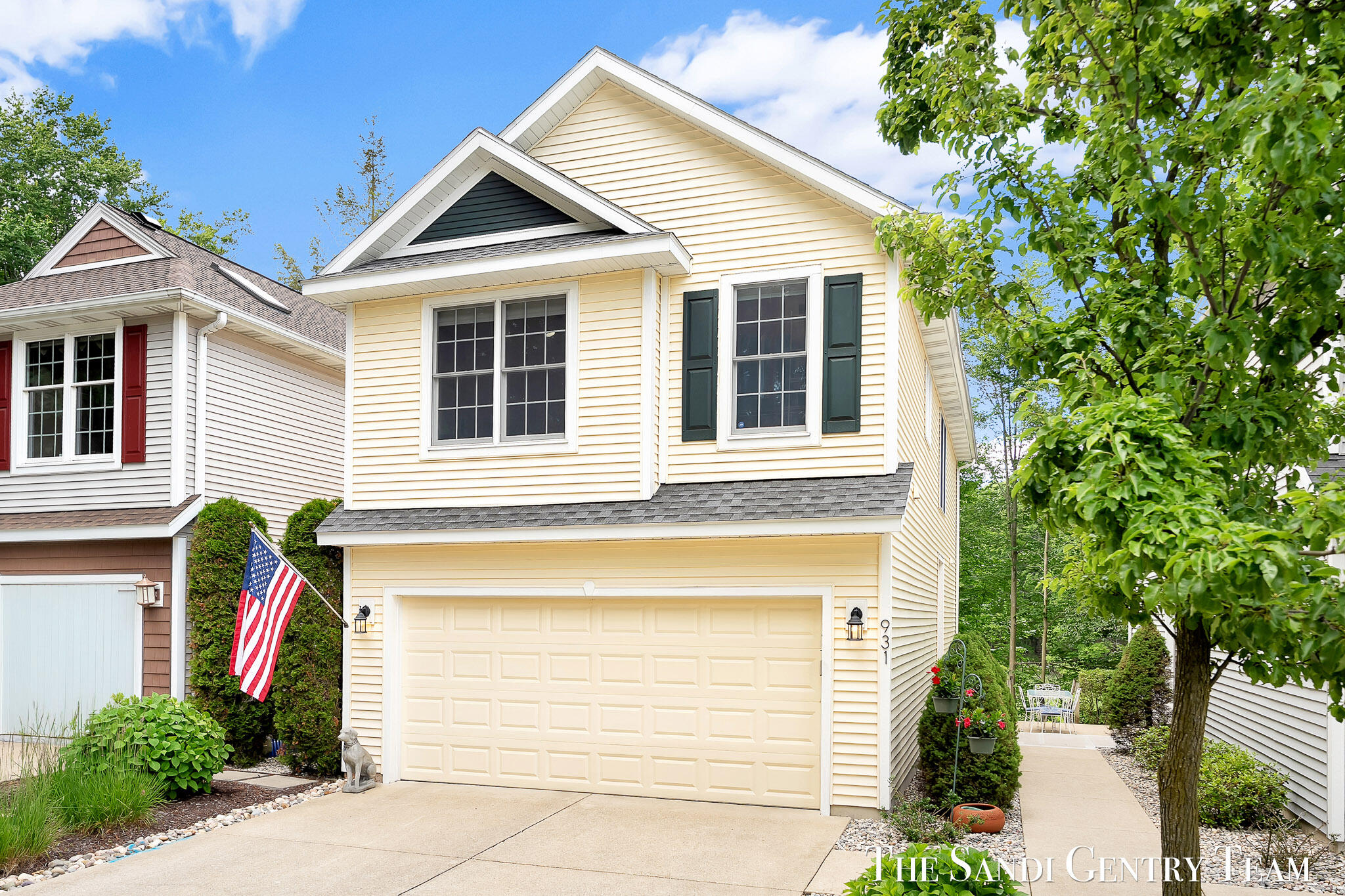 Pale yellow two-story home with a two-car garage, dark green shutters, and an American flag.