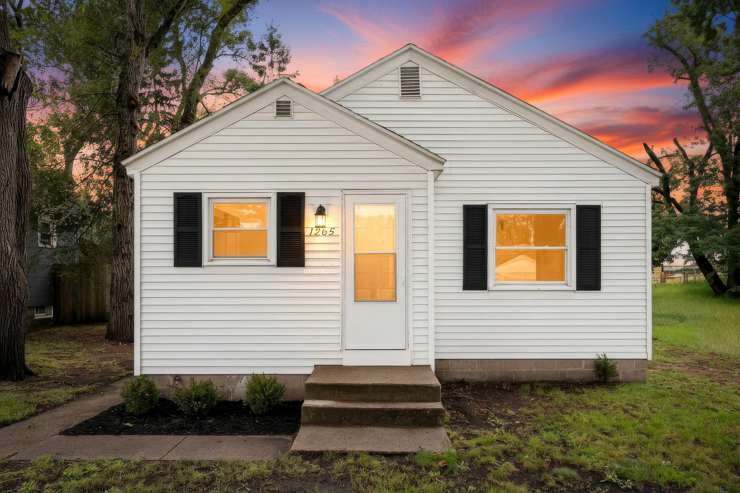 White-sided house with black shutters and glowing windows beneath a pink-orange dusk sky.