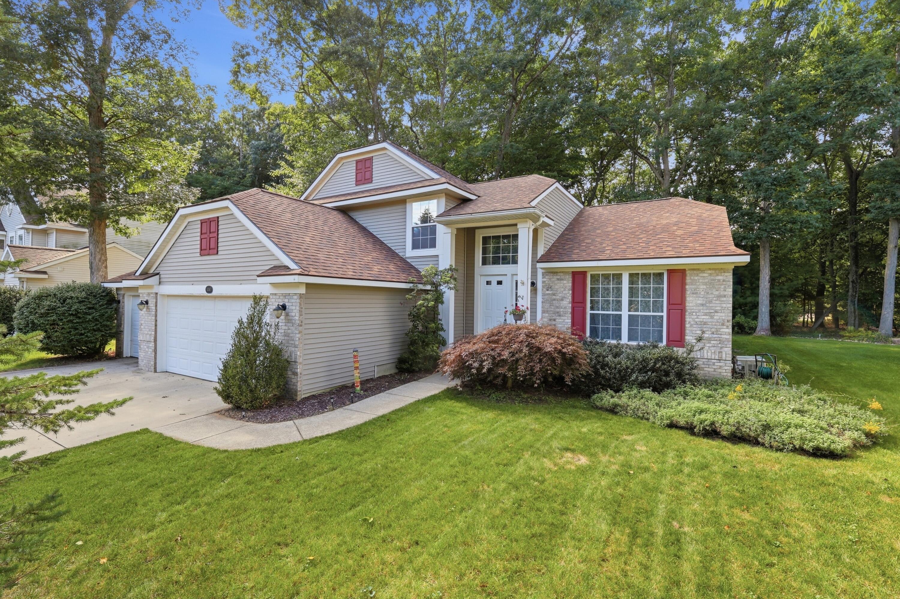 Two-story beige home with red shutters, stone accents, and an attached garage, surrounded by mature trees.