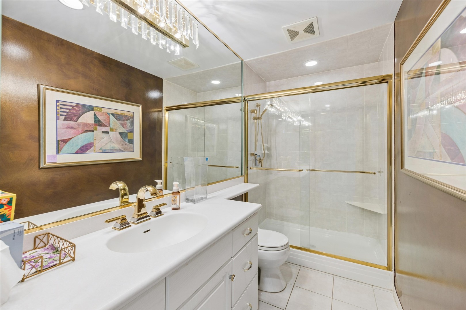 Bathroom with a long white vanity and glass shower framed in gold-tone trim.