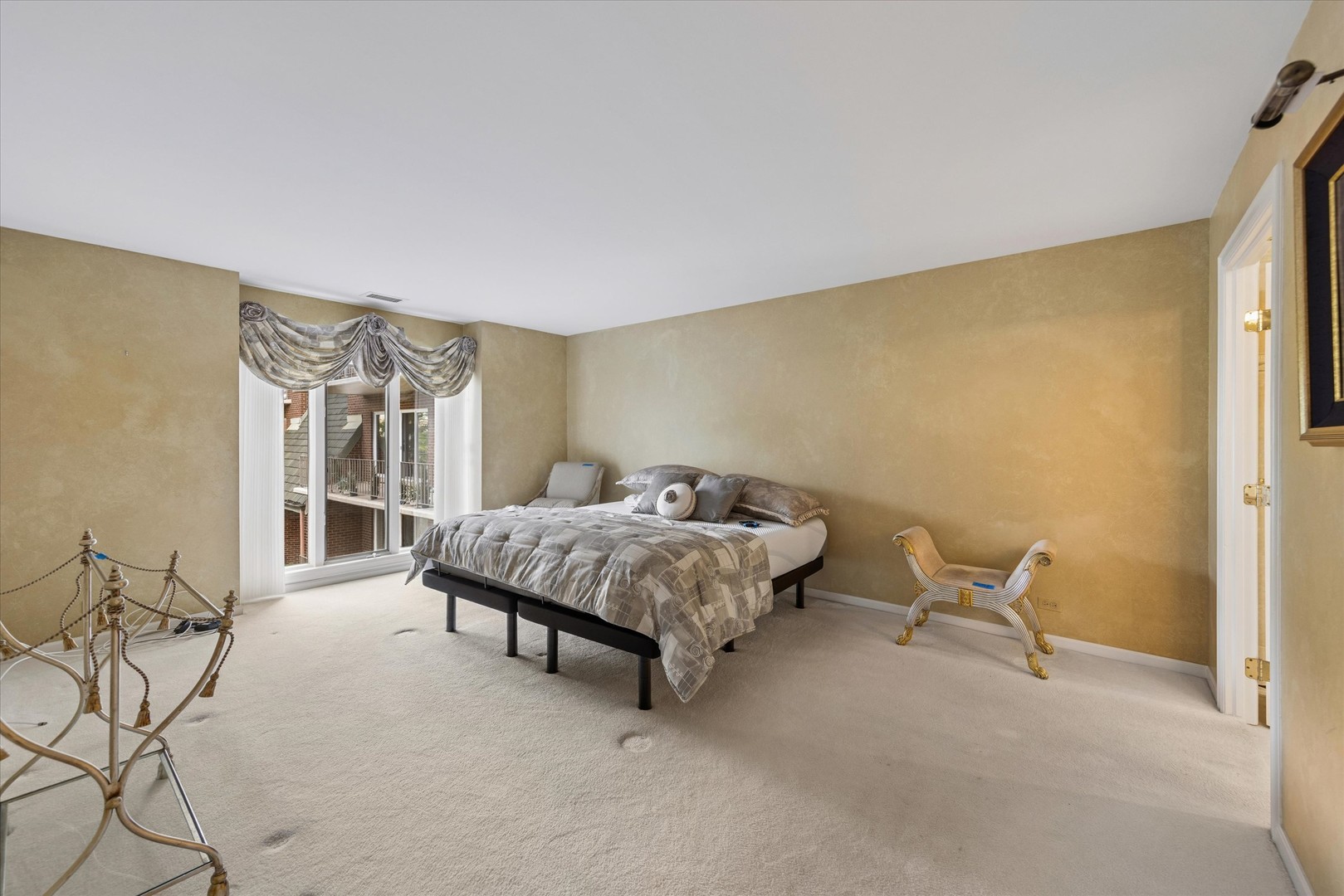 Spacious bedroom with a gray-patterned bed, beige walls, and large curtained windows.