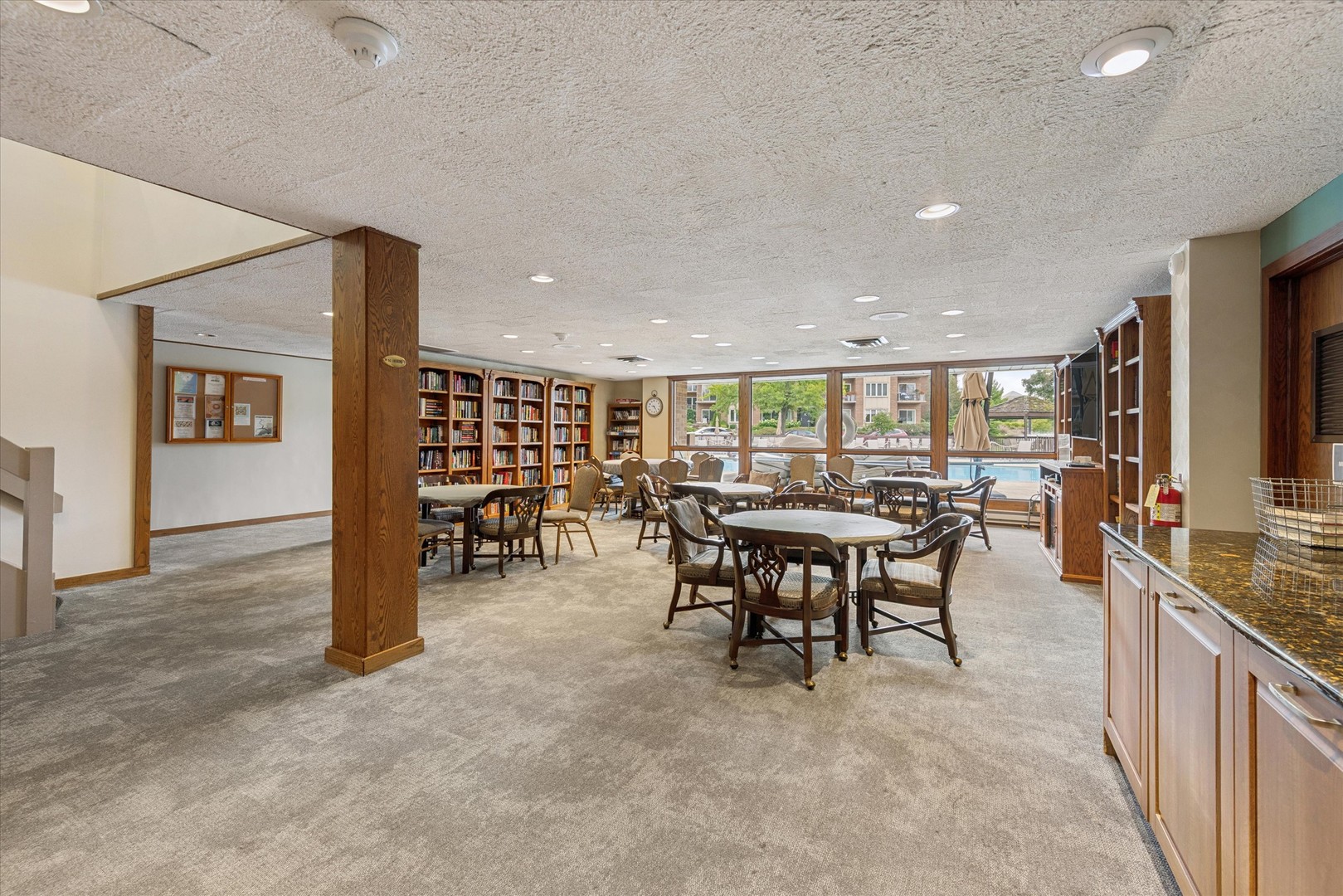Community room with round tables, bookshelves, and windows overlooking an outdoor pool.
