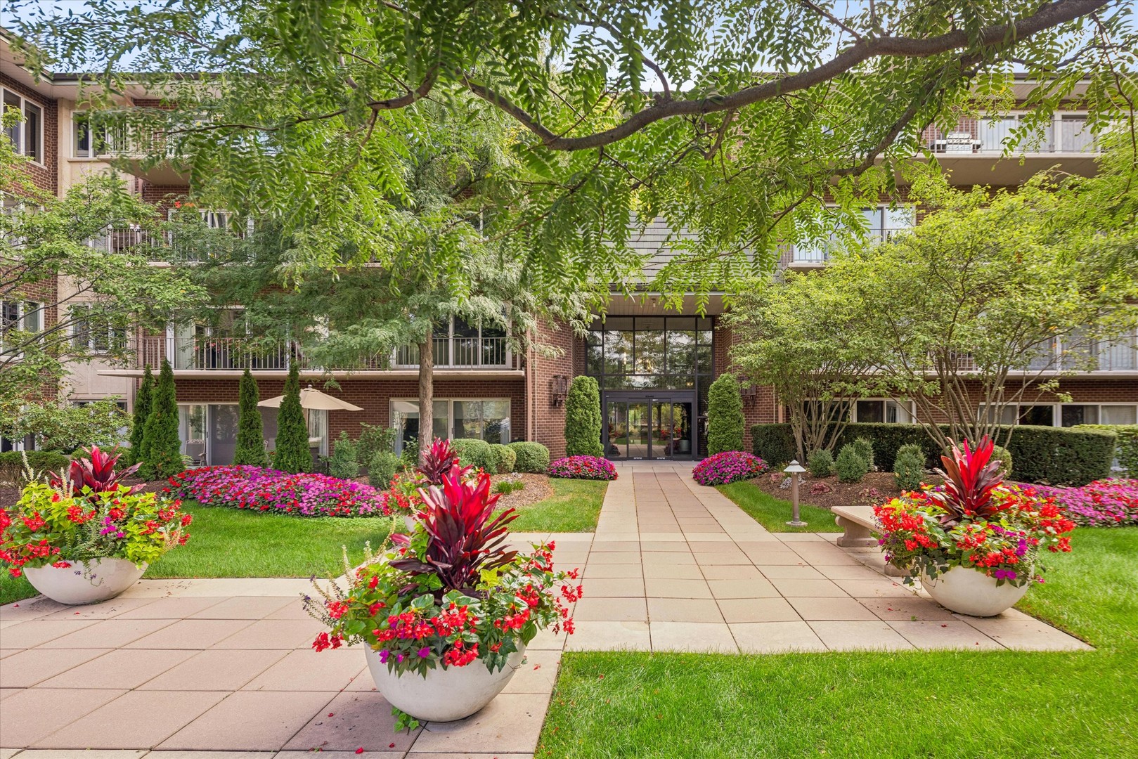 Brick condominium entrance framed by balconies, mature trees, and colorful flower beds.