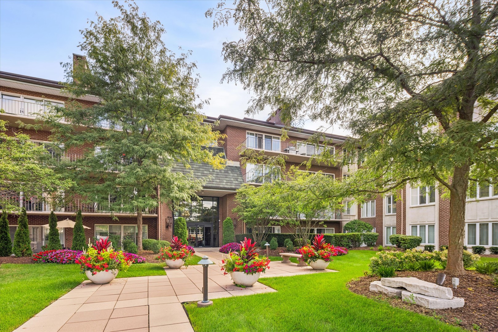 Brick condominium building with balconies behind a landscaped courtyard and flower-filled planters.