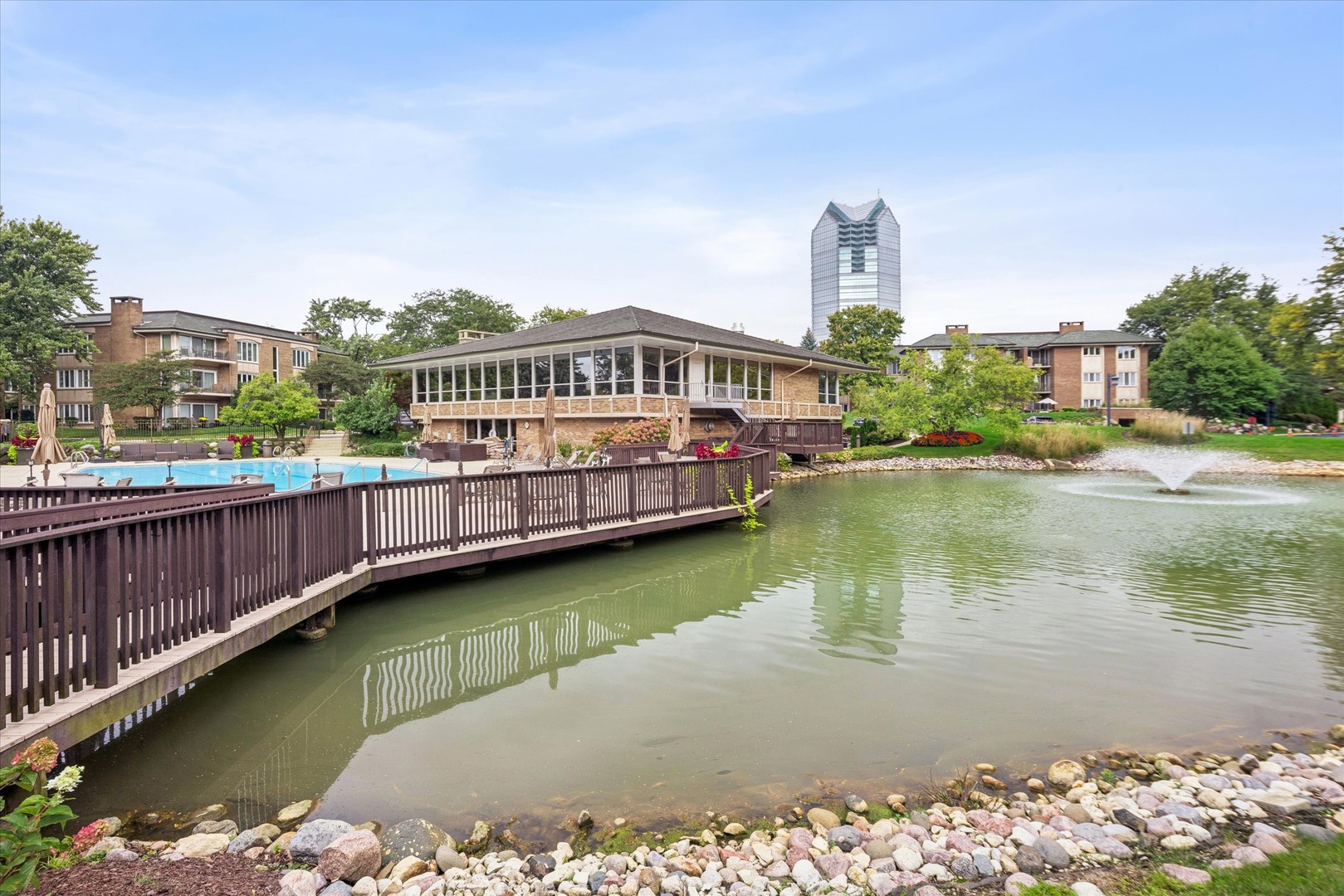 Condominium clubhouse and pool beside a landscaped pond with a fountain and distant high-rise.