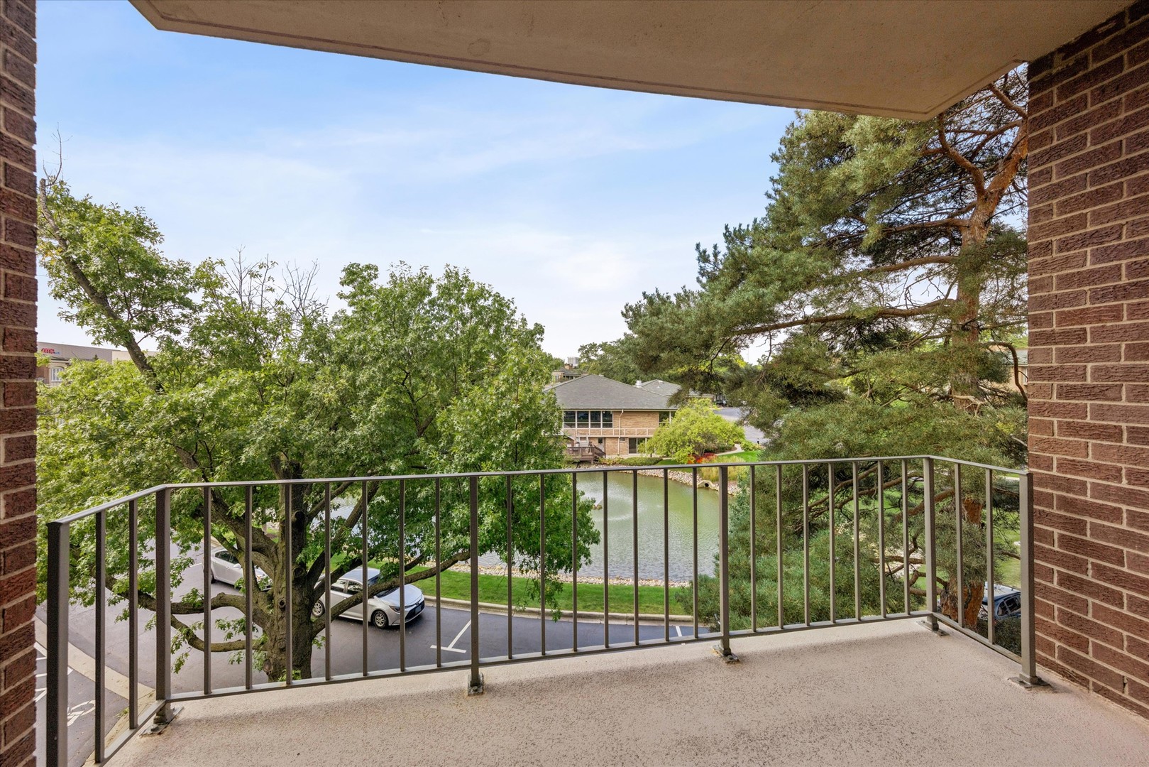 Covered balcony overlooking a pond, leafy trees, and a parking area.