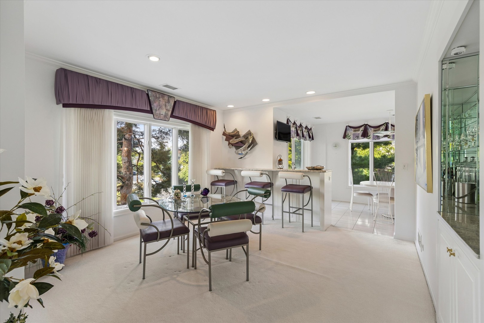 Dining area with a glass-top table, breakfast bar, and large windows overlooking trees.