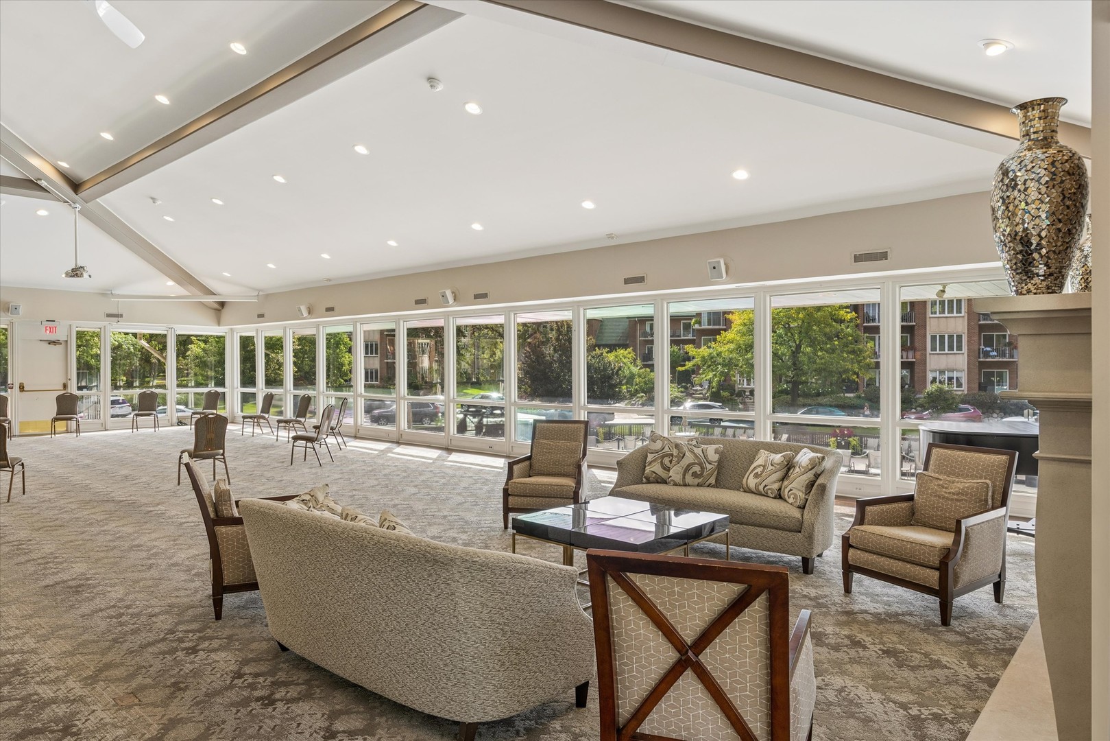 Community lounge with seating, vaulted ceiling, and floor-to-ceiling windows overlooking trees and neighboring buildings.
