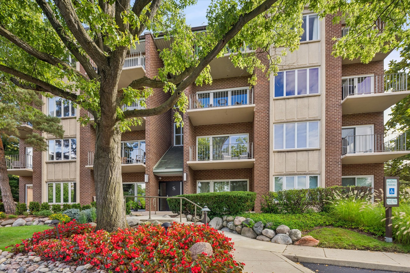 Brick condominium exterior with balconies, a large shade tree, and colorful flower beds along the entrance walkway.