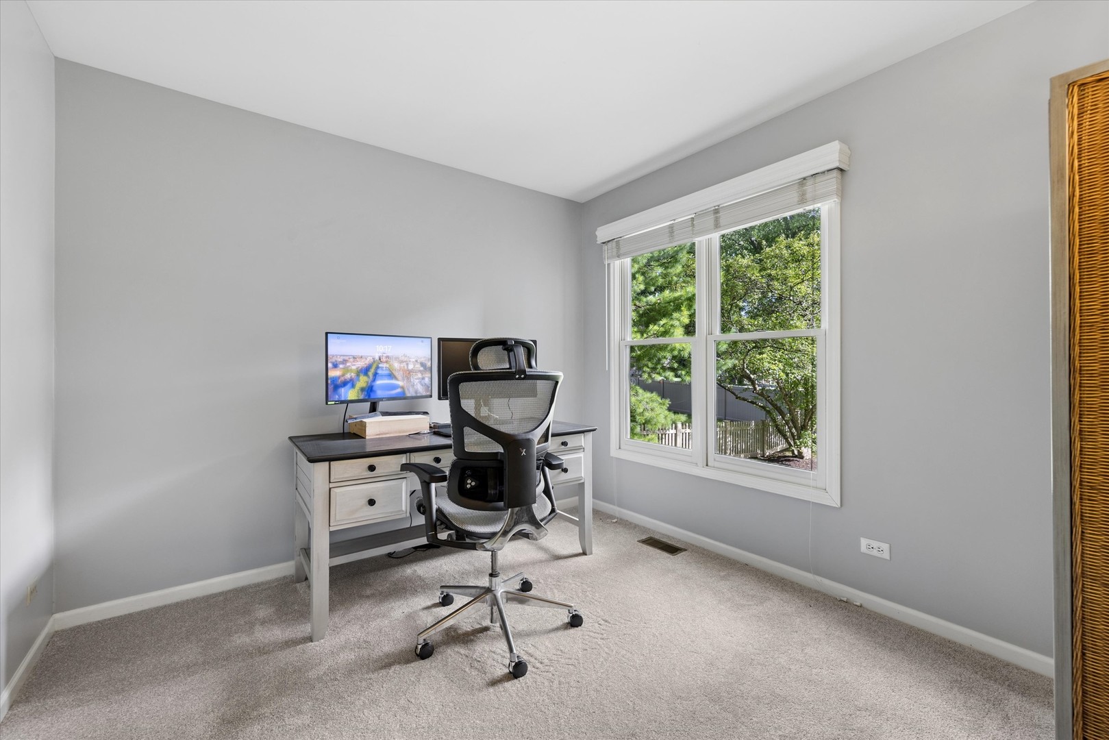 Home office with a desk and ergonomic chair beside a large window overlooking leafy trees.