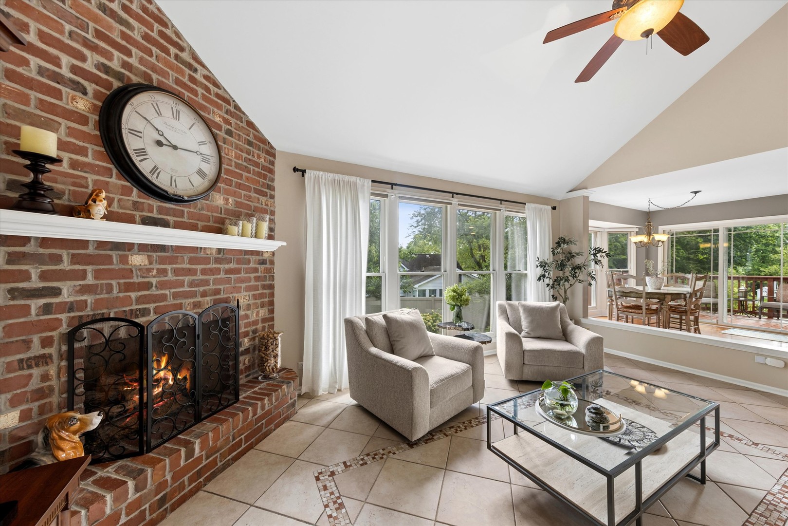 Living room with a curved brick fireplace, pale armchairs, and large windows beside a dining area.