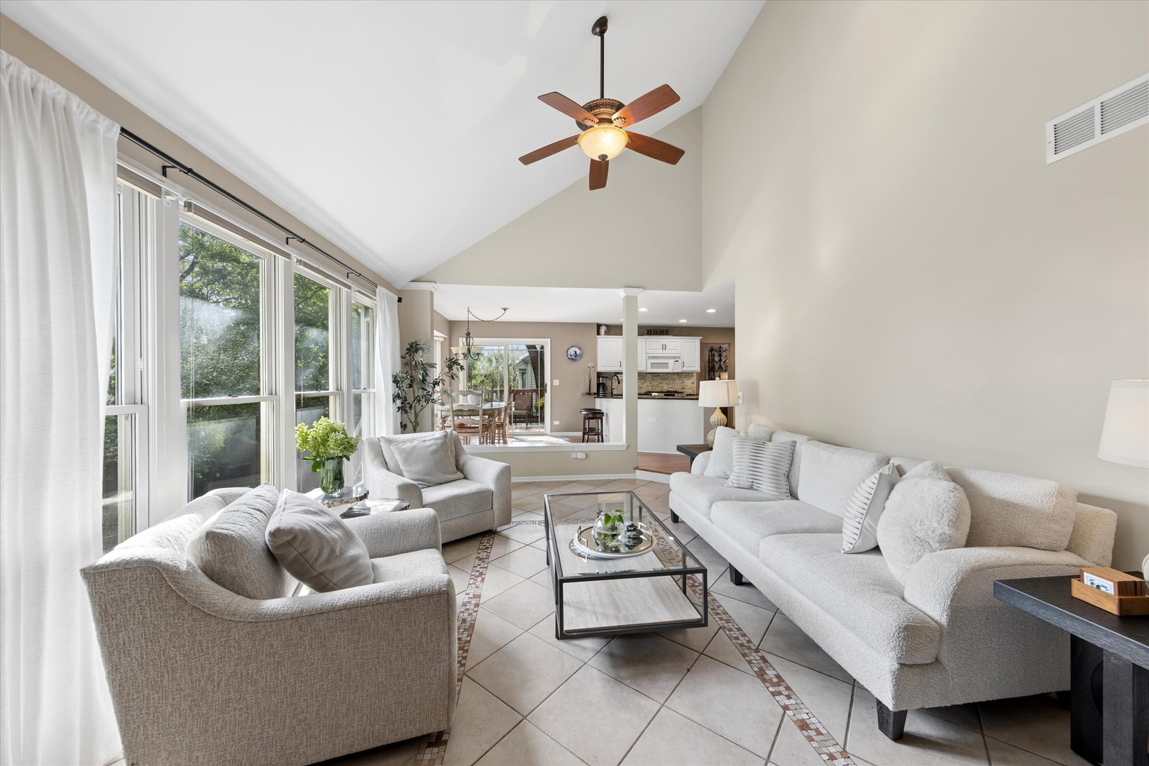 Staged living room with vaulted ceiling, large windows, white sofas, and tiled floor.