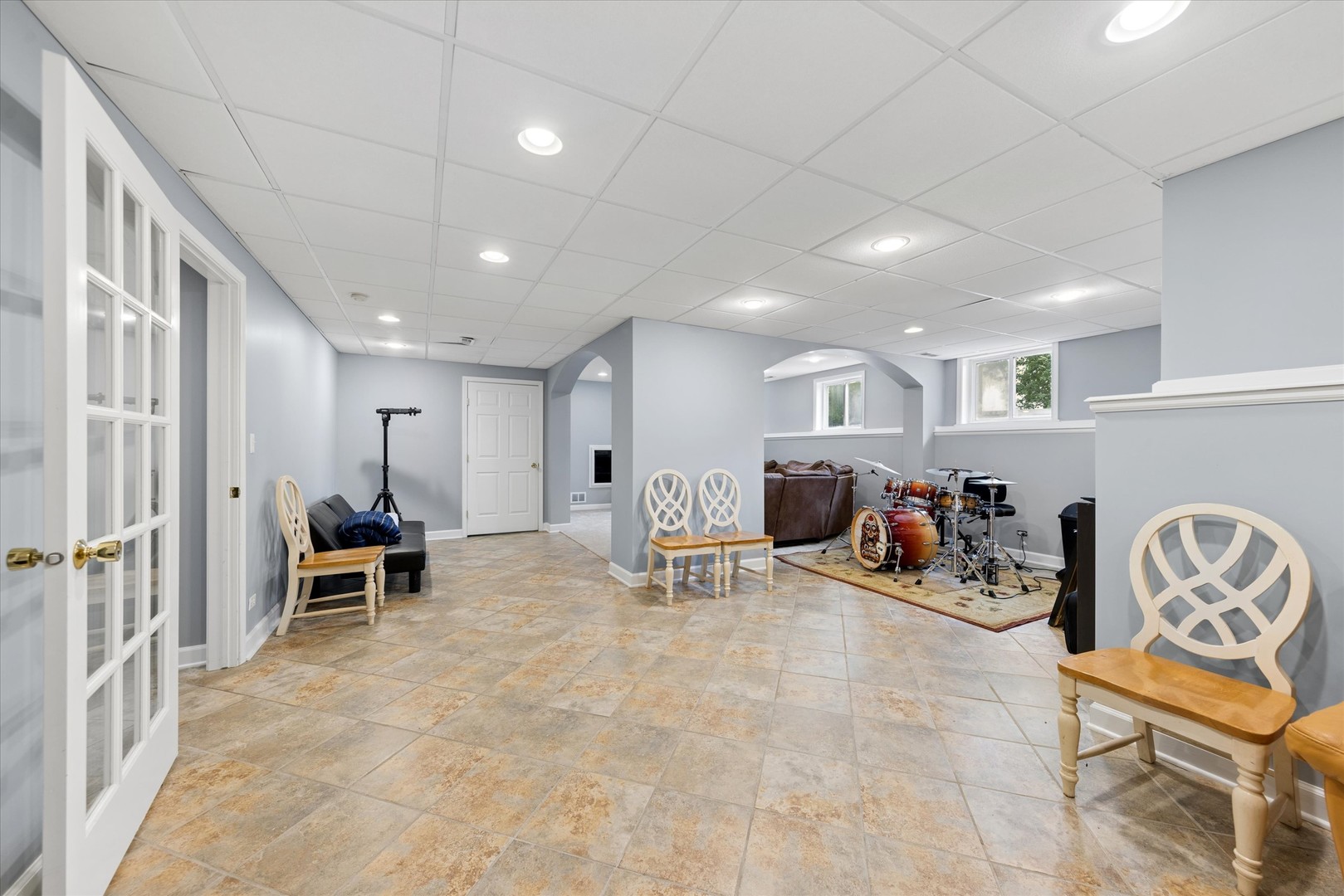 Furnished recreation room with a drum kit, brown sectional sofa, tiled floor, and arched openings.