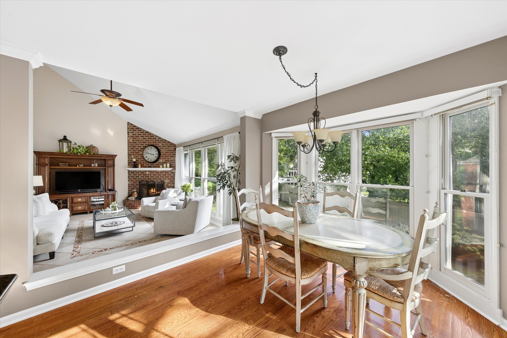 Dining room with an oval table by large windows, adjoining a living room with a brick fireplace.