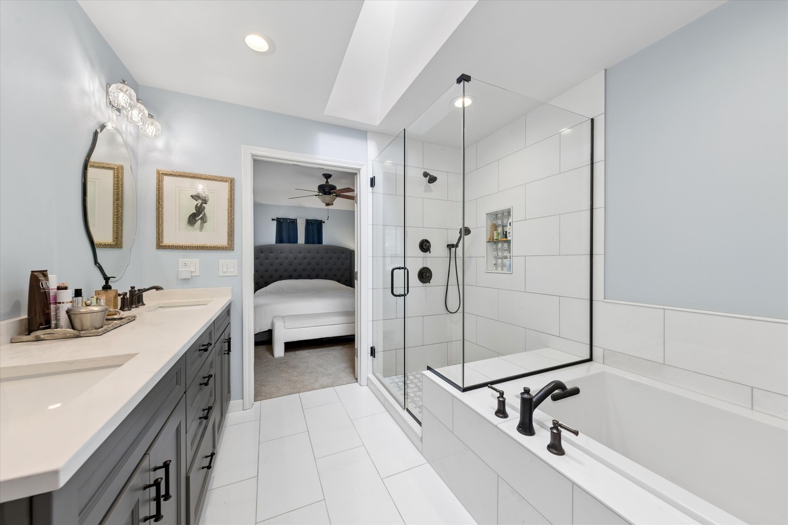 Renovated bathroom with a glass shower, white bathtub, and gray vanity; bedroom visible through the doorway.