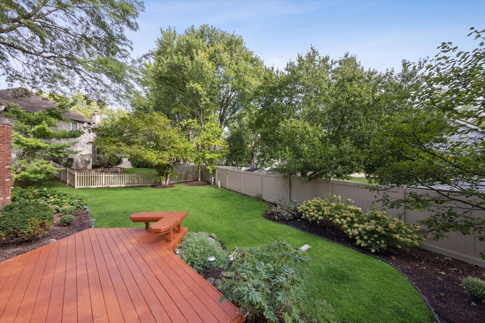 Fenced backyard with a green lawn, mature trees, and a reddish wood deck with built-in bench.