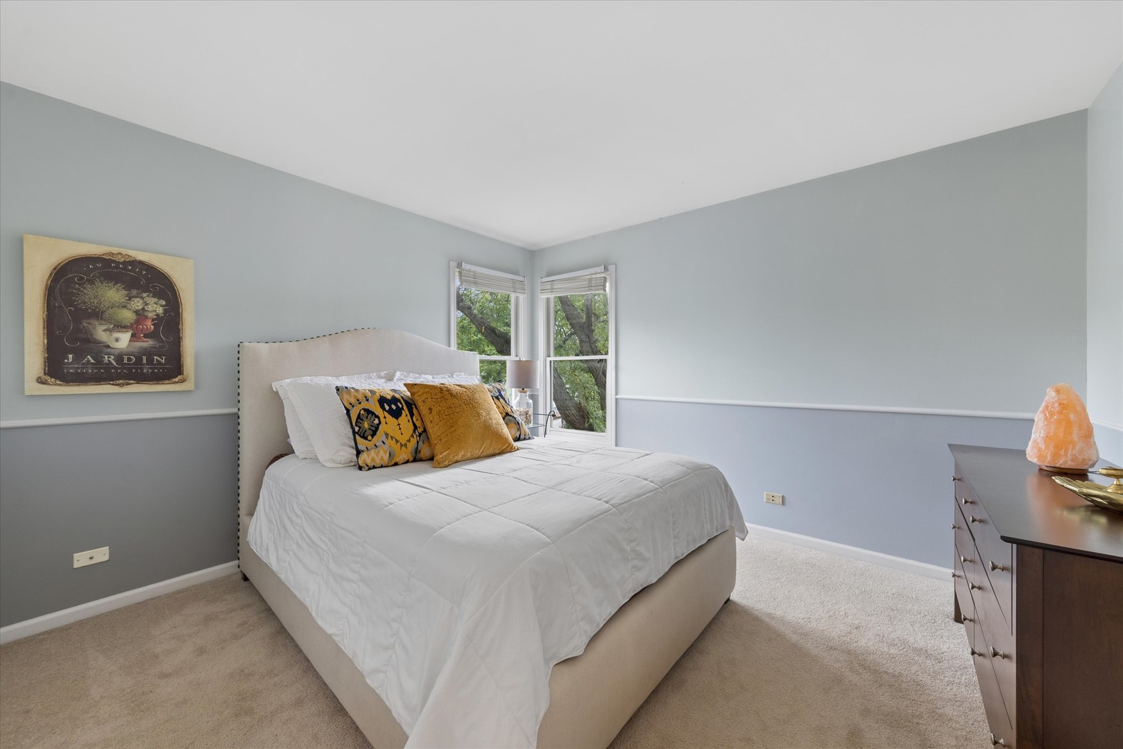Staged bedroom with a white bed, mustard accent pillows, and corner windows overlooking leafy trees.