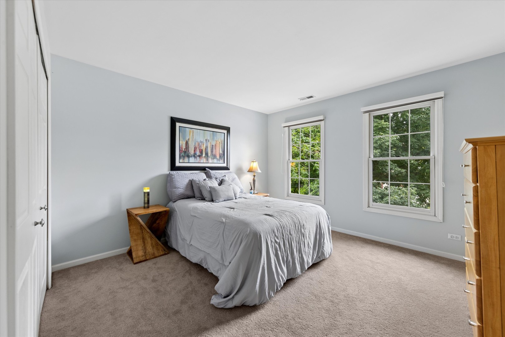 Furnished bedroom with a gray bed, beige carpet, and two windows overlooking leafy trees.