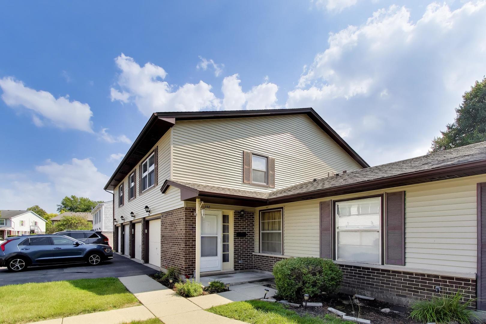 Two-story attached home with beige siding, a brick lower facade, and a landscaped entrance.