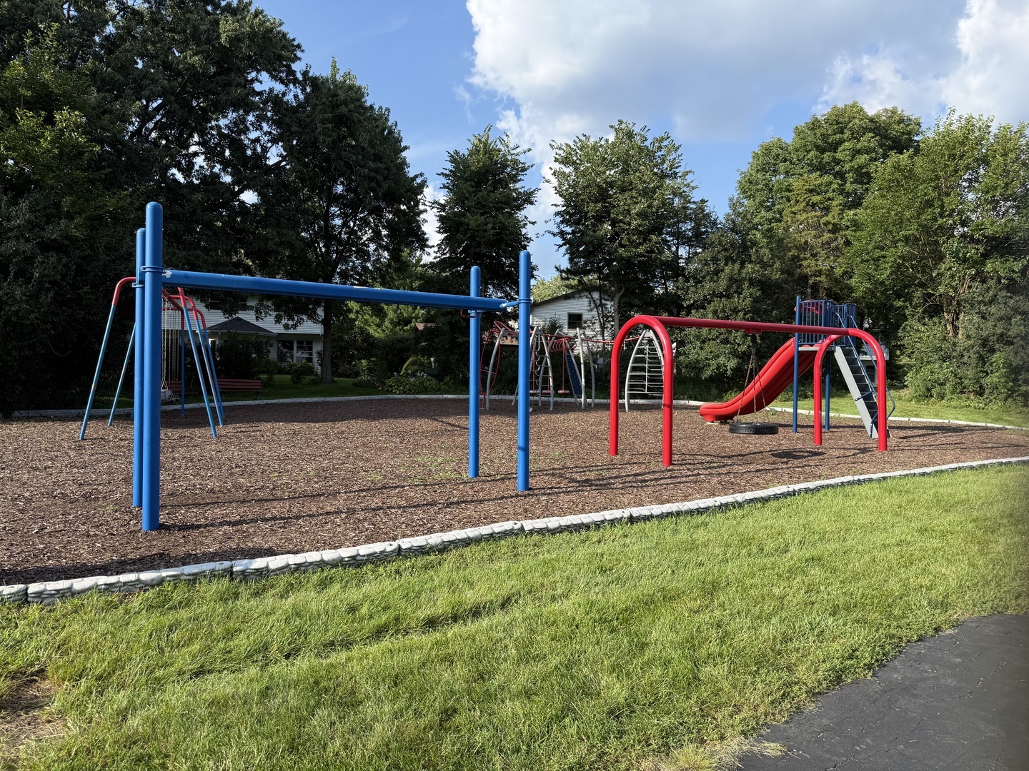 Playground with blue swings and red slides, surrounded by grass and mature trees.