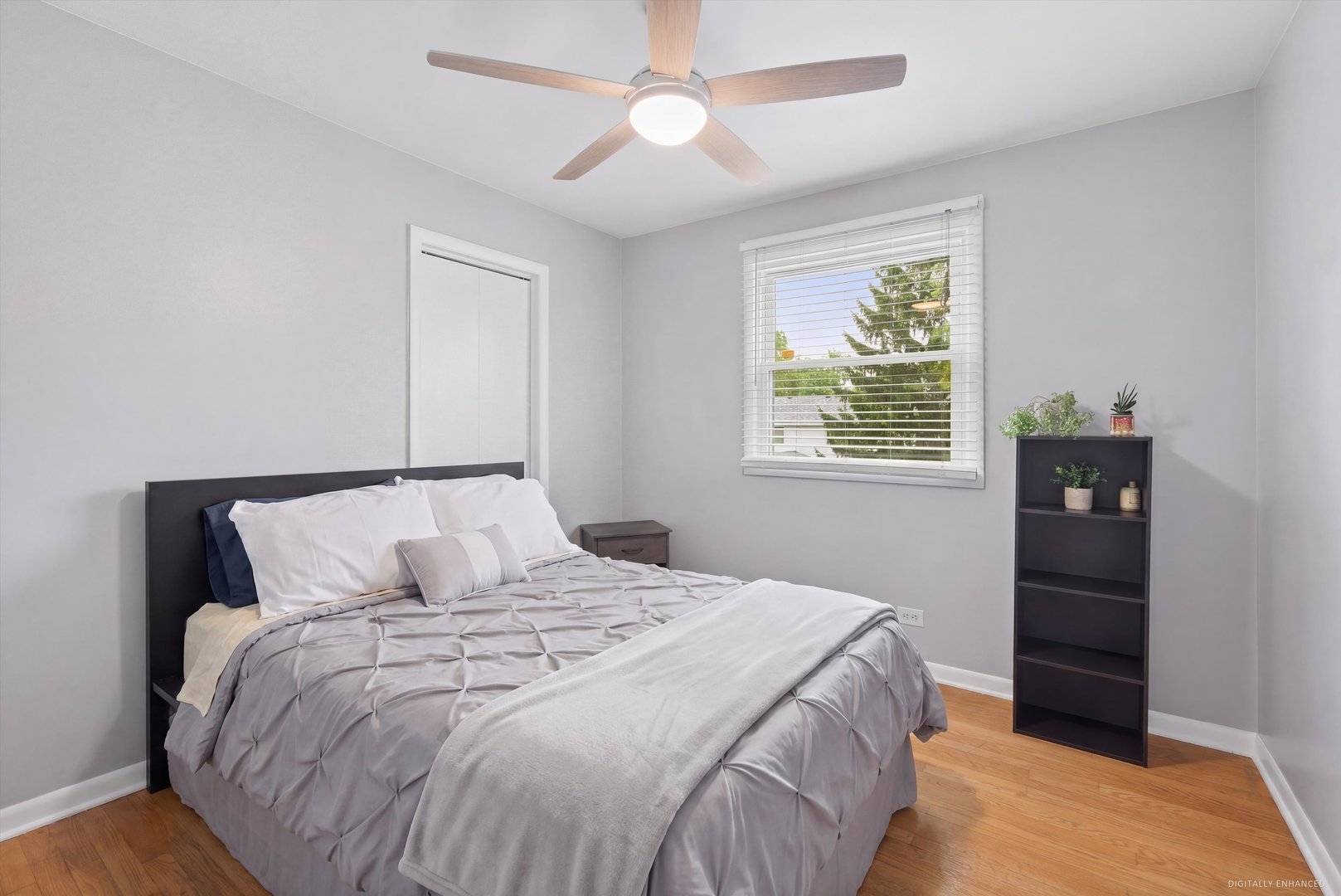 Staged bedroom with a gray bed, ceiling fan, window with blinds, and wood flooring.