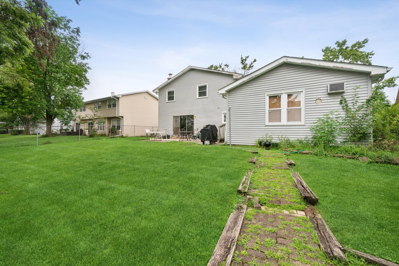 Rear of a light gray two-story house with a broad lawn, patio furniture, and a worn brick path.