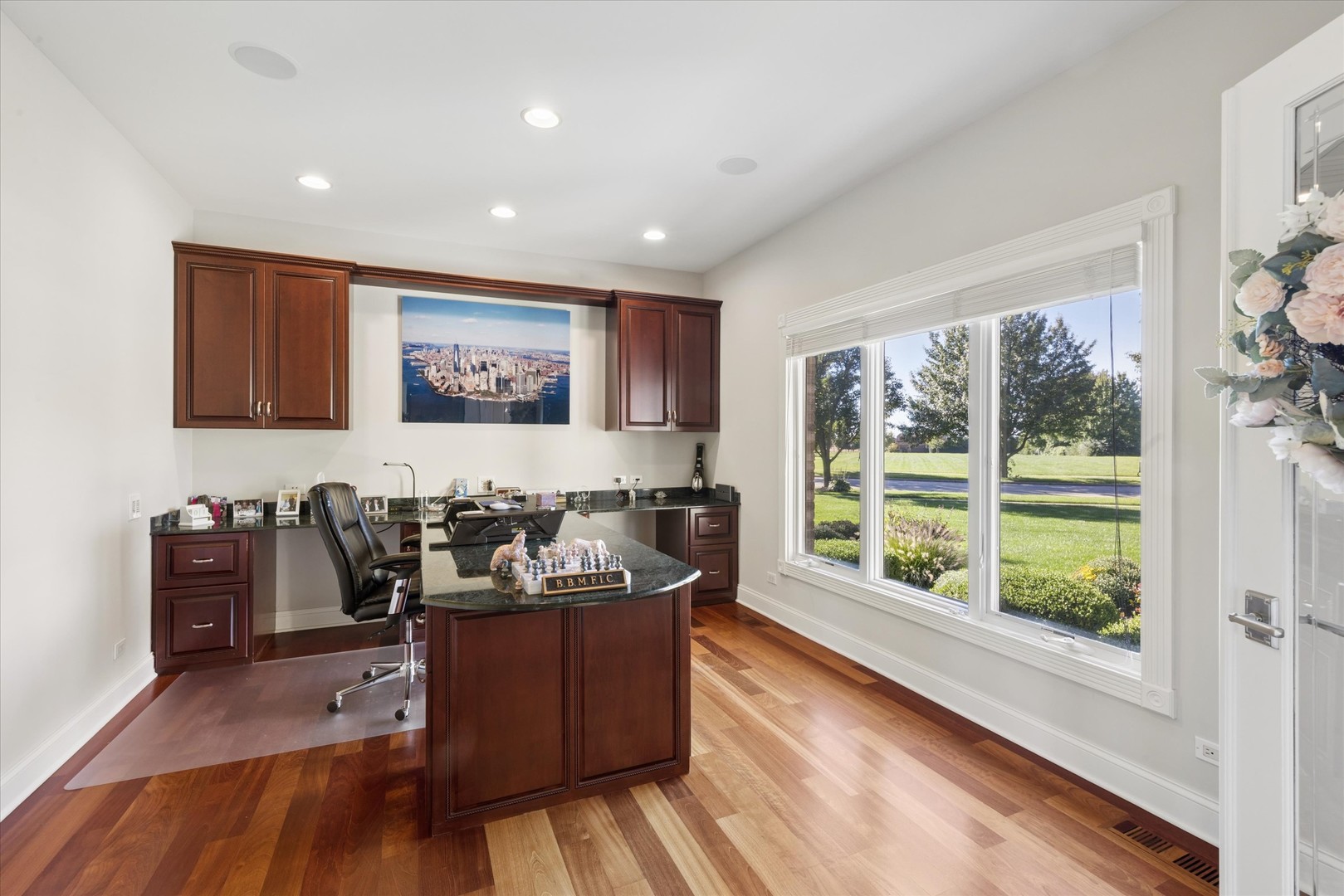 Home office with dark wood cabinetry, a central desk, hardwood floors, and large windows overlooking a lawn.