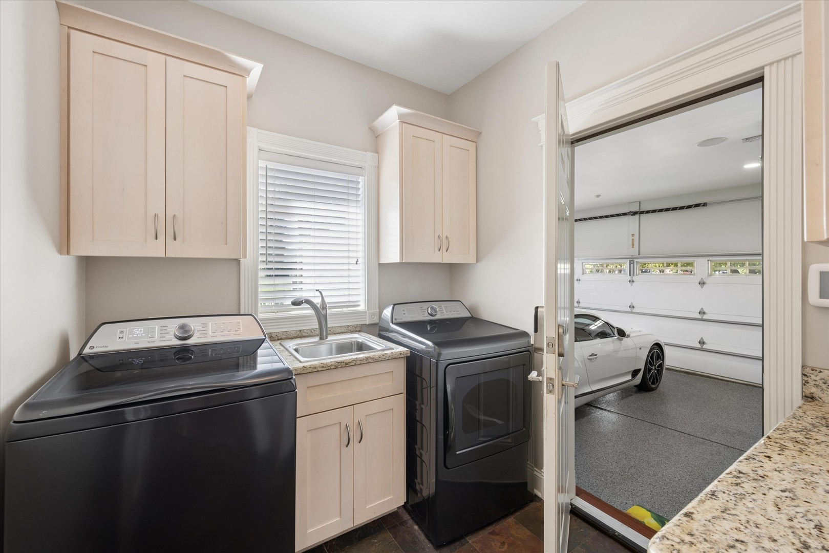Laundry room with dark washer and dryer, utility sink, and an open doorway to a garage with a white car.