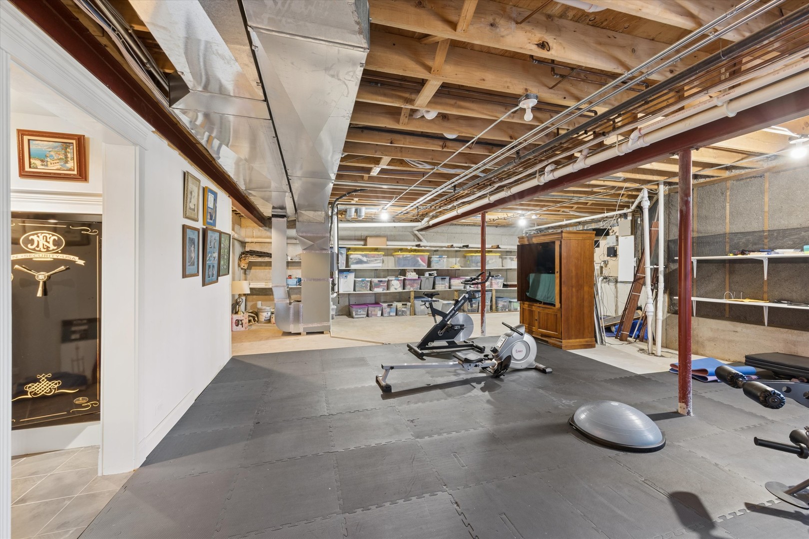 Basement gym with exercise bikes, rubber floor mats, and storage shelves beneath exposed ceiling joists.