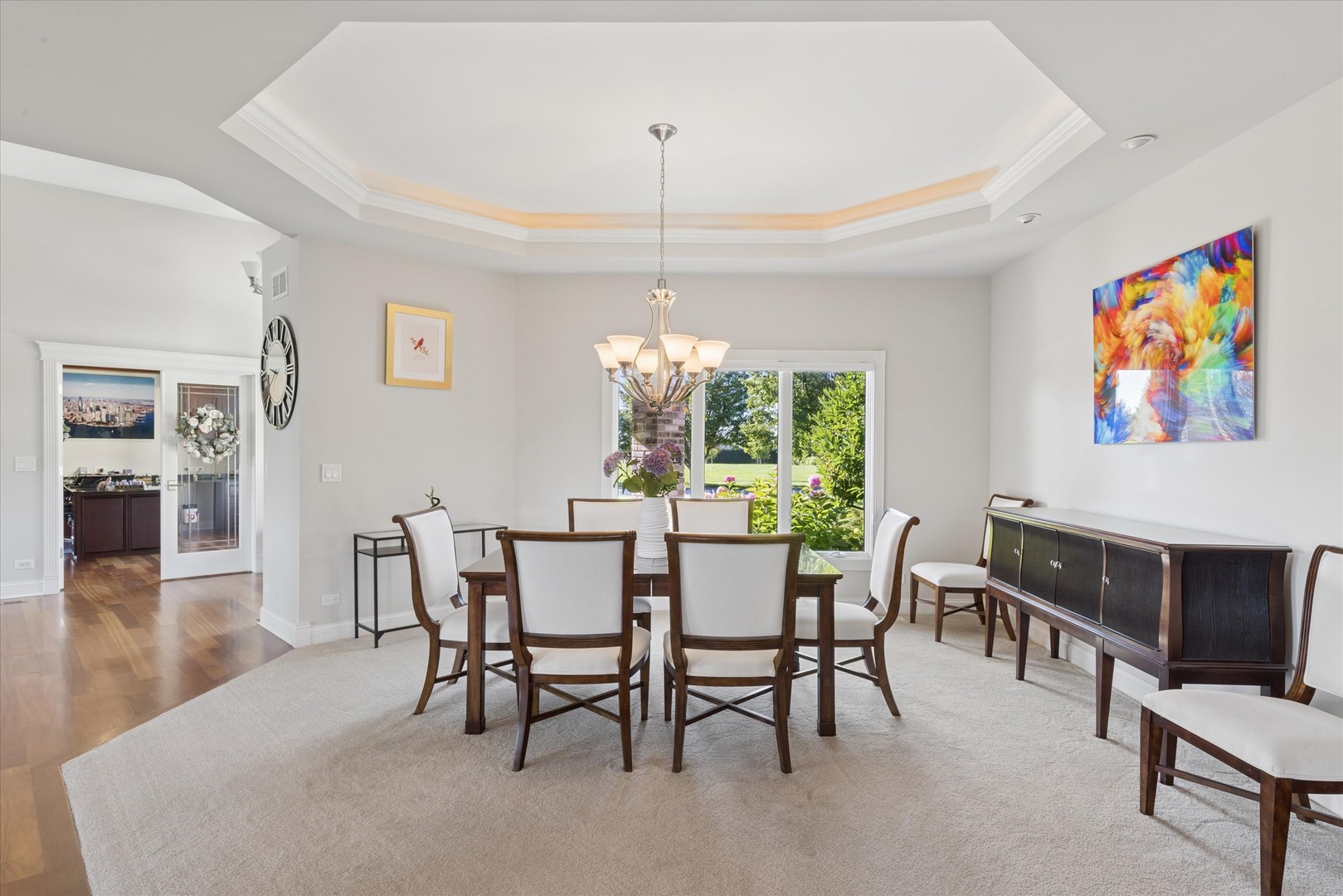 Dining room with a wood table, chandelier, tray ceiling, and garden view through a large window.