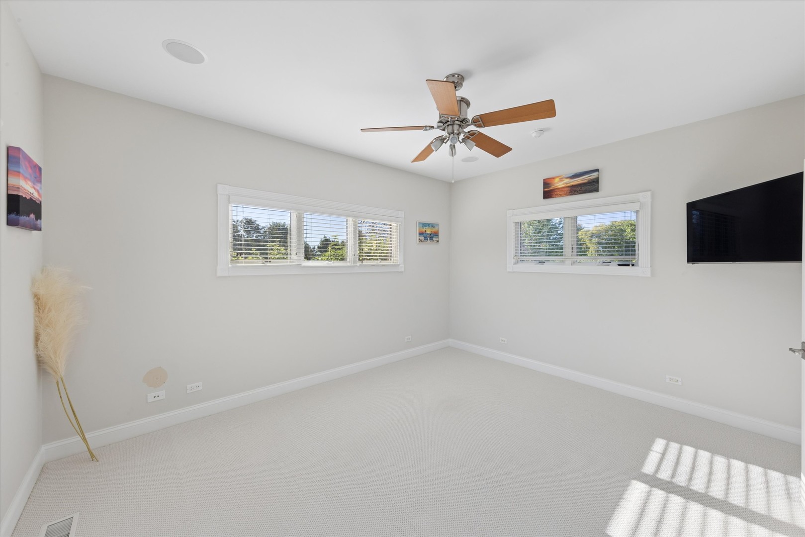 Bright carpeted bedroom with two blind-covered windows, a ceiling fan, and a wall-mounted TV.
