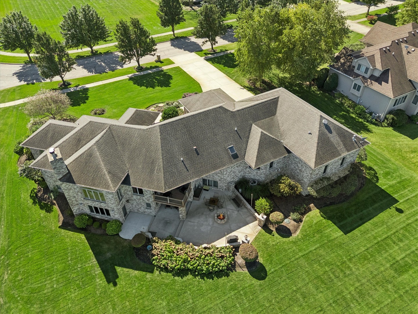 Aerial view of a stone-sided home with a landscaped lawn and rear patio with a fire pit.