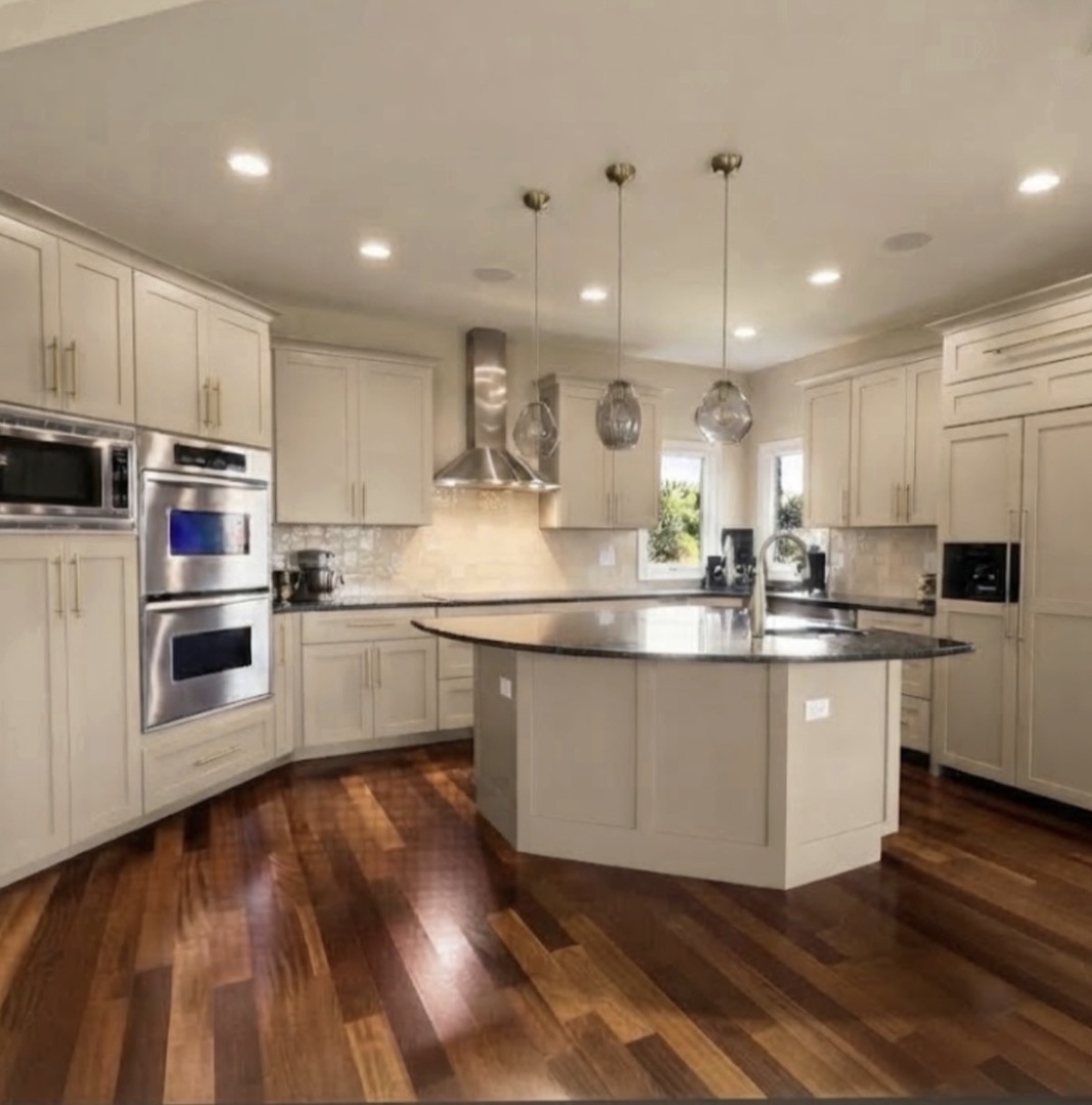 Kitchen with a large dark stone island, cream cabinets, stainless steel double ovens, and glossy hardwood floors.