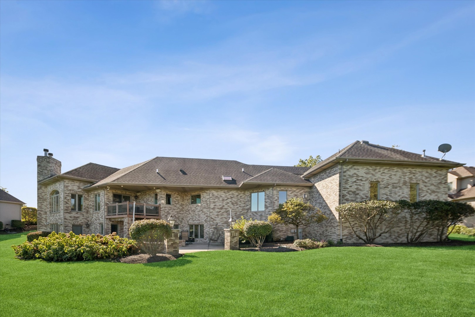 Brick home’s rear exterior with a raised deck, patio swing, landscaped shrubs, and broad lawn.