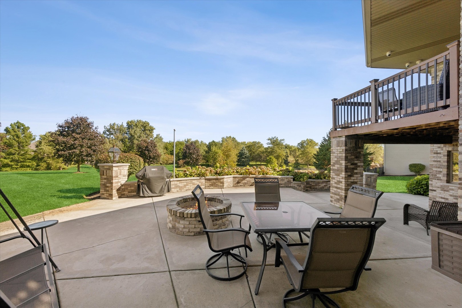 Patio with a stone fire pit, outdoor dining set, and raised deck overlooking a lawn and trees.