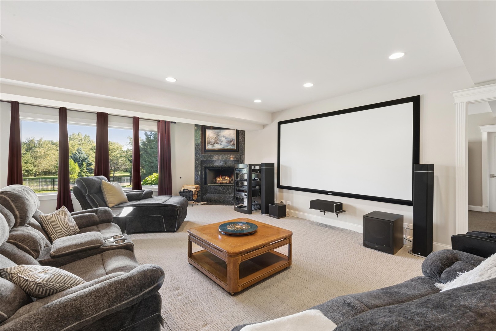 Furnished home theater with a large projection screen, fireplace, gray sofas, and windows overlooking trees.