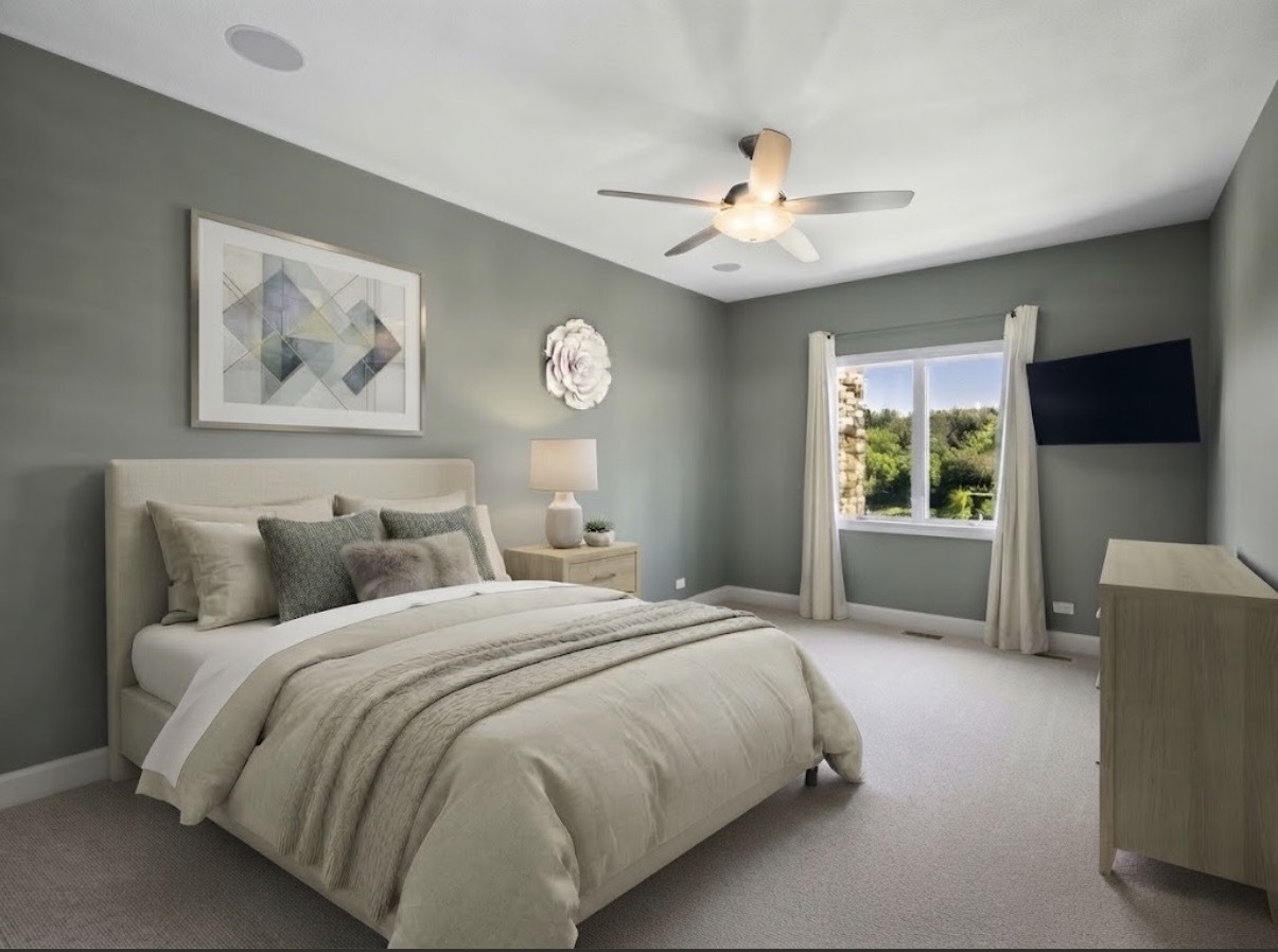 Staged bedroom with a cream bed, ceiling fan, and window overlooking trees.
