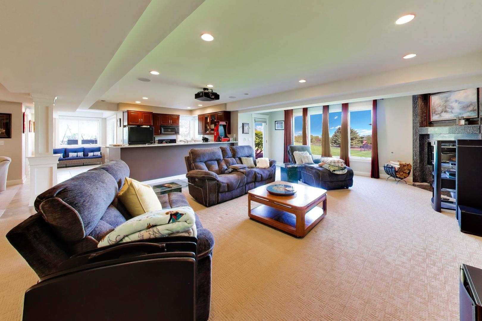 Furnished living room open to the kitchen, with a fireplace and large windows overlooking a lawn.