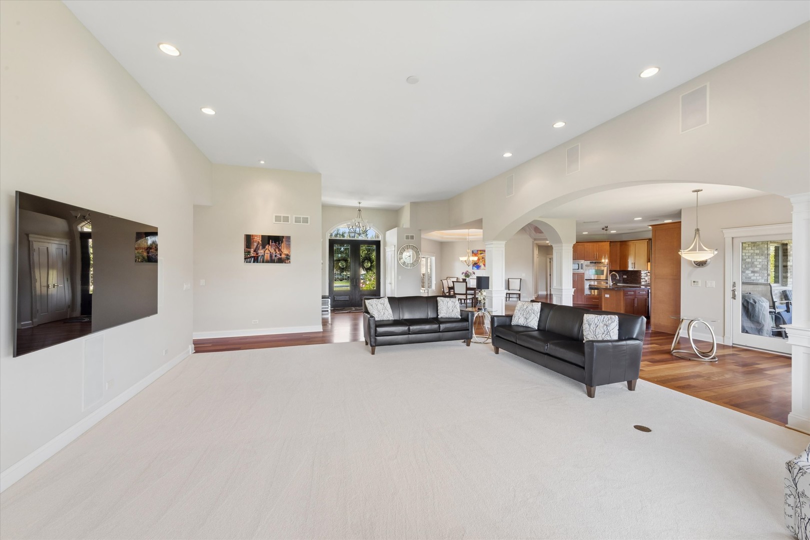 Open-plan living room with black leather sofas, a large cream carpet, and an adjoining kitchen and dining area.