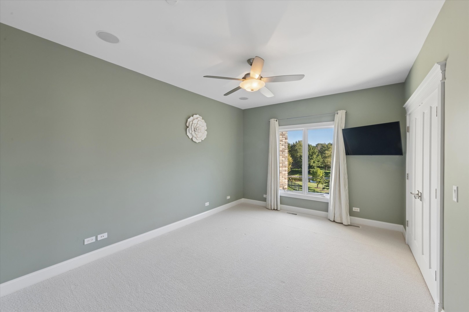 Empty bedroom with sage walls, light carpet, ceiling fan, and a window overlooking trees.