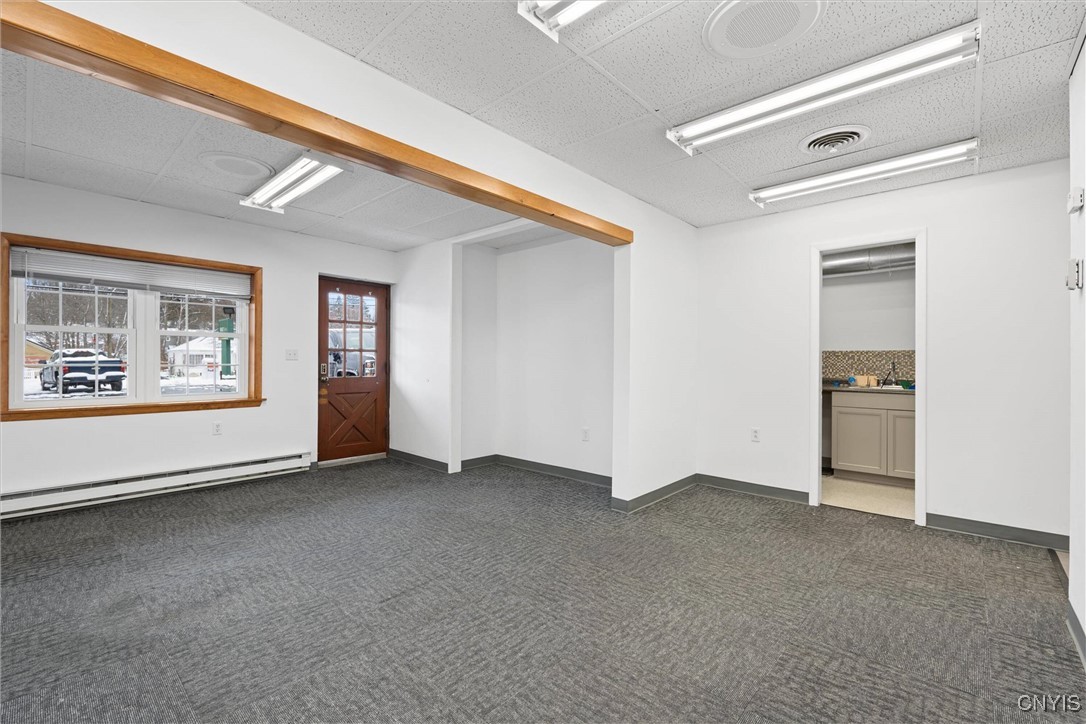Vacant office interior with gray carpet, a wood-trimmed window, and a doorway to a kitchenette.