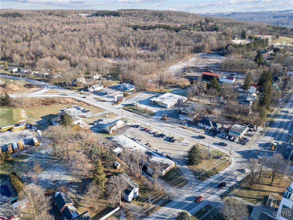 Aerial view of commercial buildings at a road intersection, with wooded hills and patches of snow.