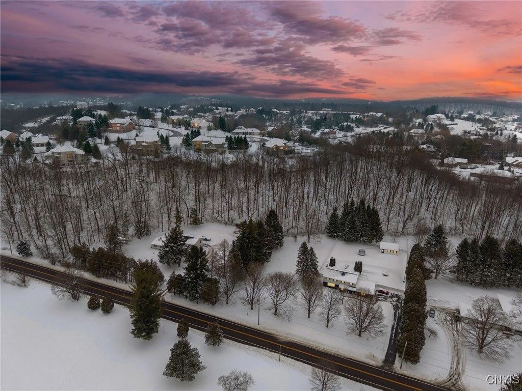 Snow-covered home and grounds beside a road, viewed aerially at dusk with woods and neighborhood beyond.