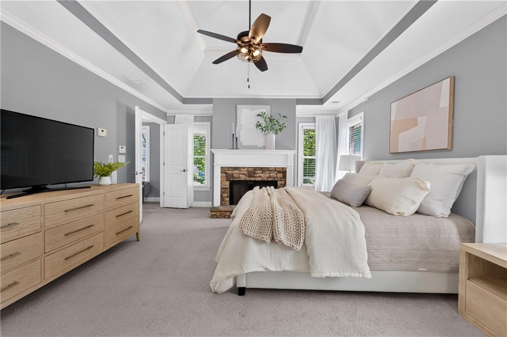 Bedroom with a white bed, tray ceiling, stone fireplace, and wooden dresser with television.